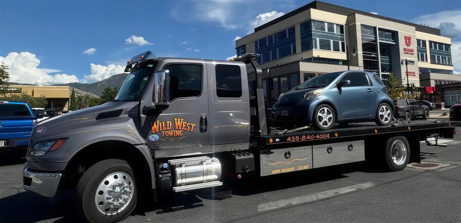 Wild West Towing flatbed carrying a small blue compact car past a modern office building on a sunny day