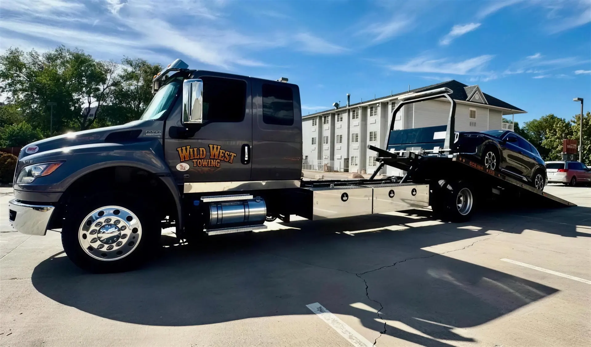 Wild West Towing grey rollback with its bed tilted down, loading a dark SUV in a parking lot in the late afternoon sun